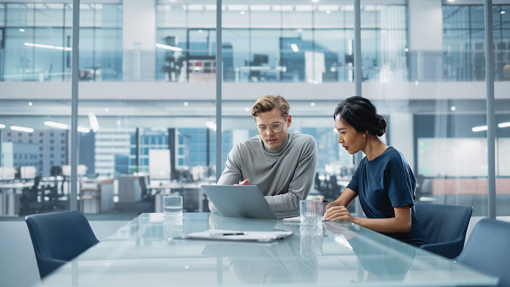 Two people sitting in a modern office viewing a laptop computer screen